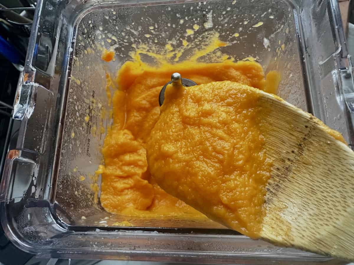 Bright orange pumpkin puree in a blender, with a wooden spatula scooping some out. The mixture appears smooth and thick, showing signs of recent blending.