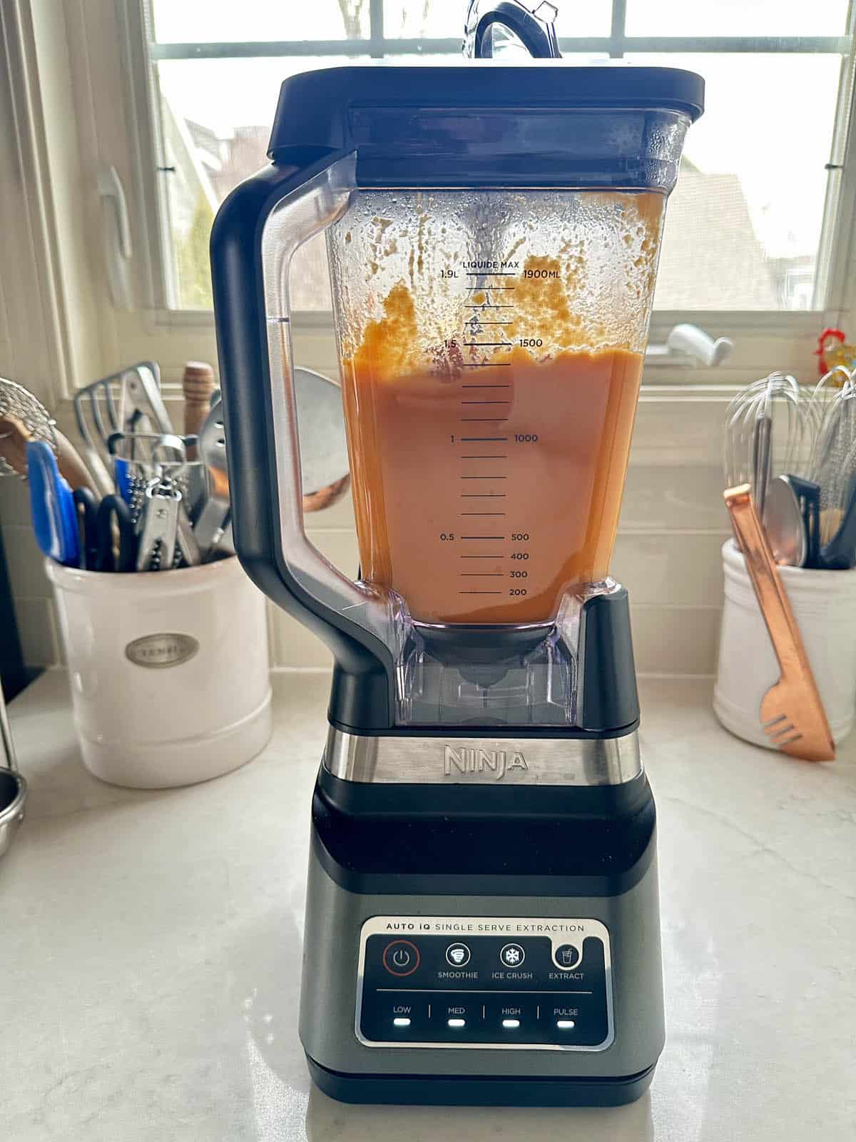 A countertop blender filled with orange soup is blending on a kitchen counter, with utensils and containers in the background near a window.