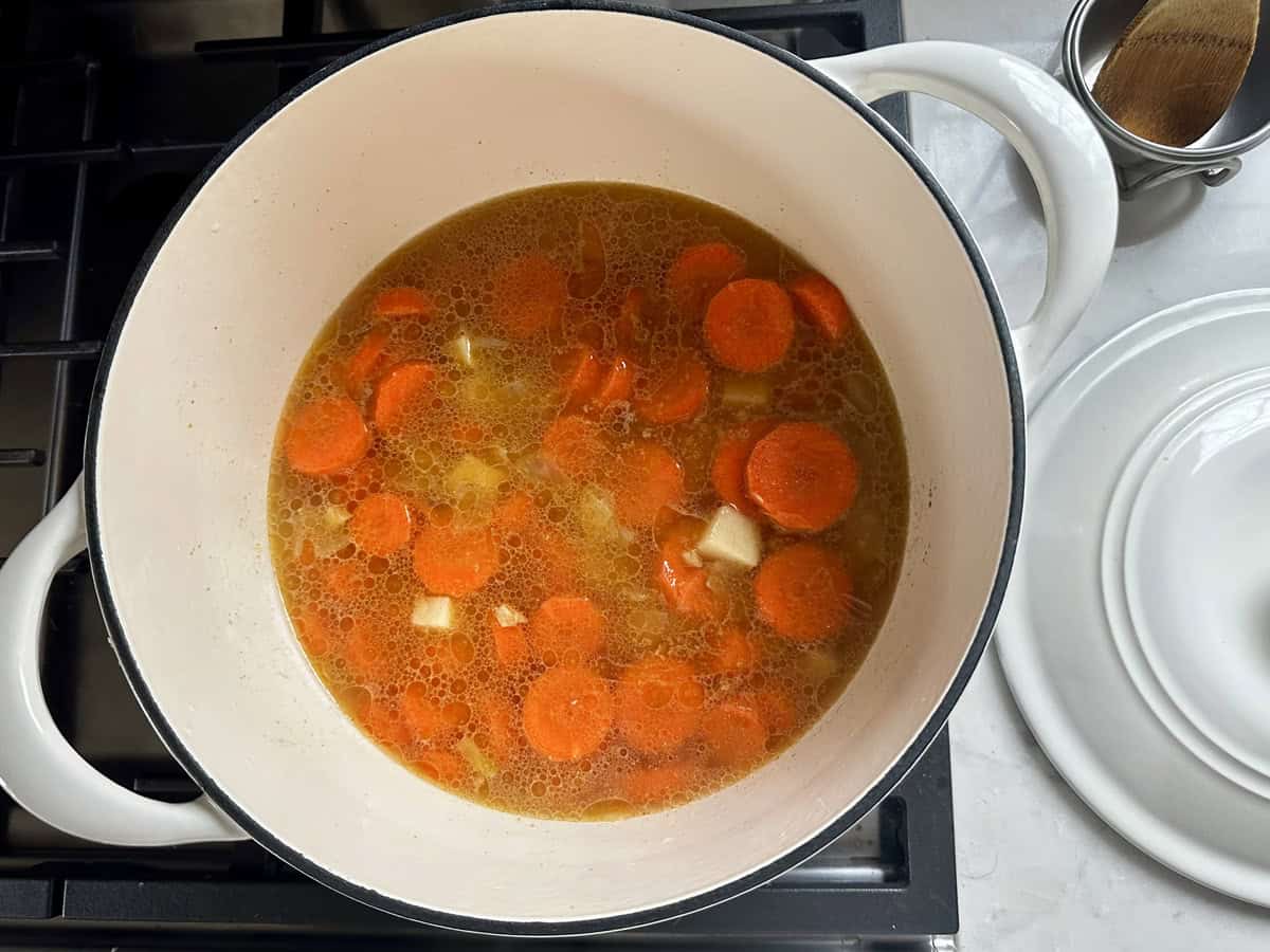 A white pot on a stove contains a simmering soup with sliced carrots and diced vegetables in a golden broth. The pot lid and a wooden spoon are on the counter nearby.