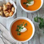 Two bowls of orange soup garnished with a dollop of cream and fresh herbs sit on a rustic wooden table. A small bowl of croutons and two spoons on a napkin are nearby.