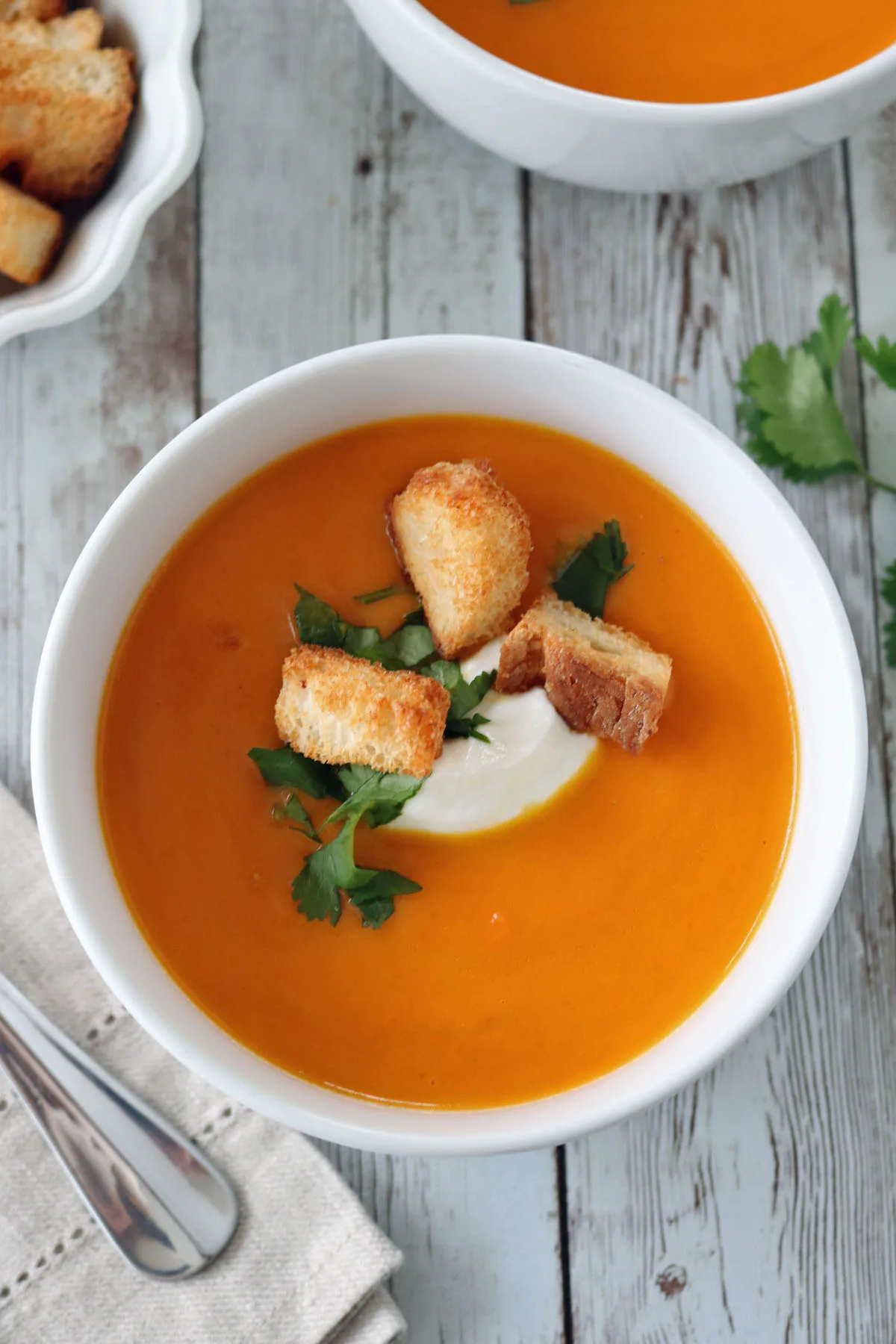 A bowl of creamy orange soup garnished with croutons, fresh cilantro, and a dollop of cream, sits on a rustic wooden table next to a napkin and spoon, with another bowl and croutons visible in the background.