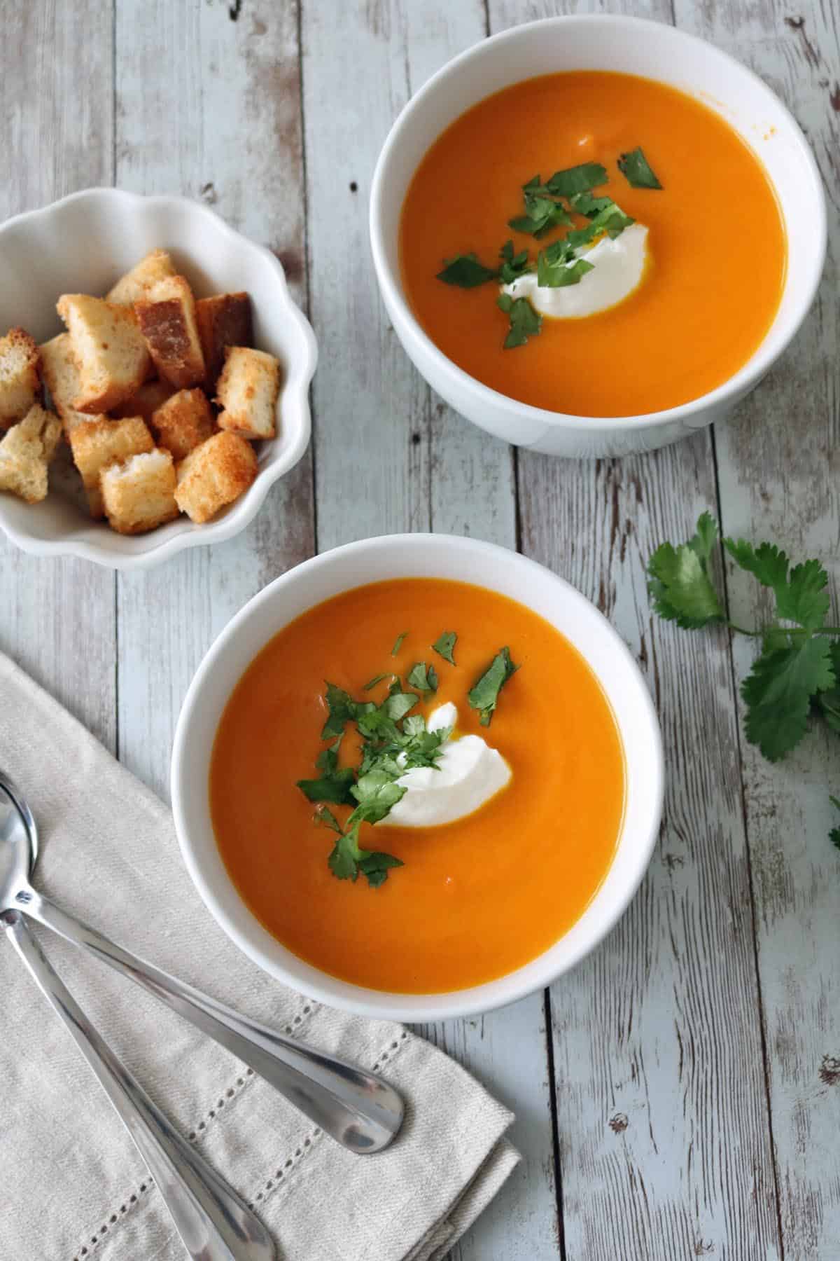 Two bowls of orange soup garnished with a dollop of cream and fresh herbs sit on a rustic wooden table. A small bowl of croutons and two spoons on a napkin are nearby.