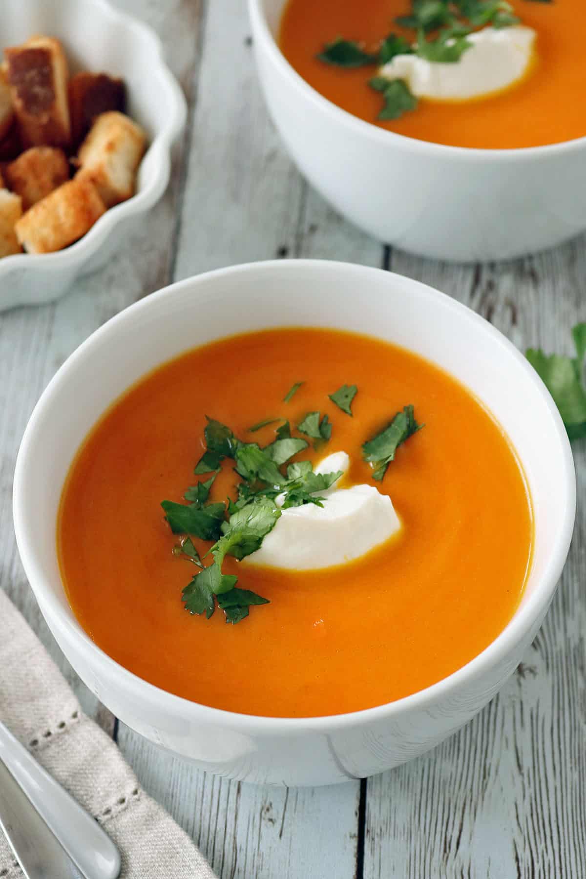 A white bowl of orange-colored soup garnished with a dollop of cream and fresh cilantro, with a side dish of croutons and another bowl of soup in the background on a rustic wooden table.