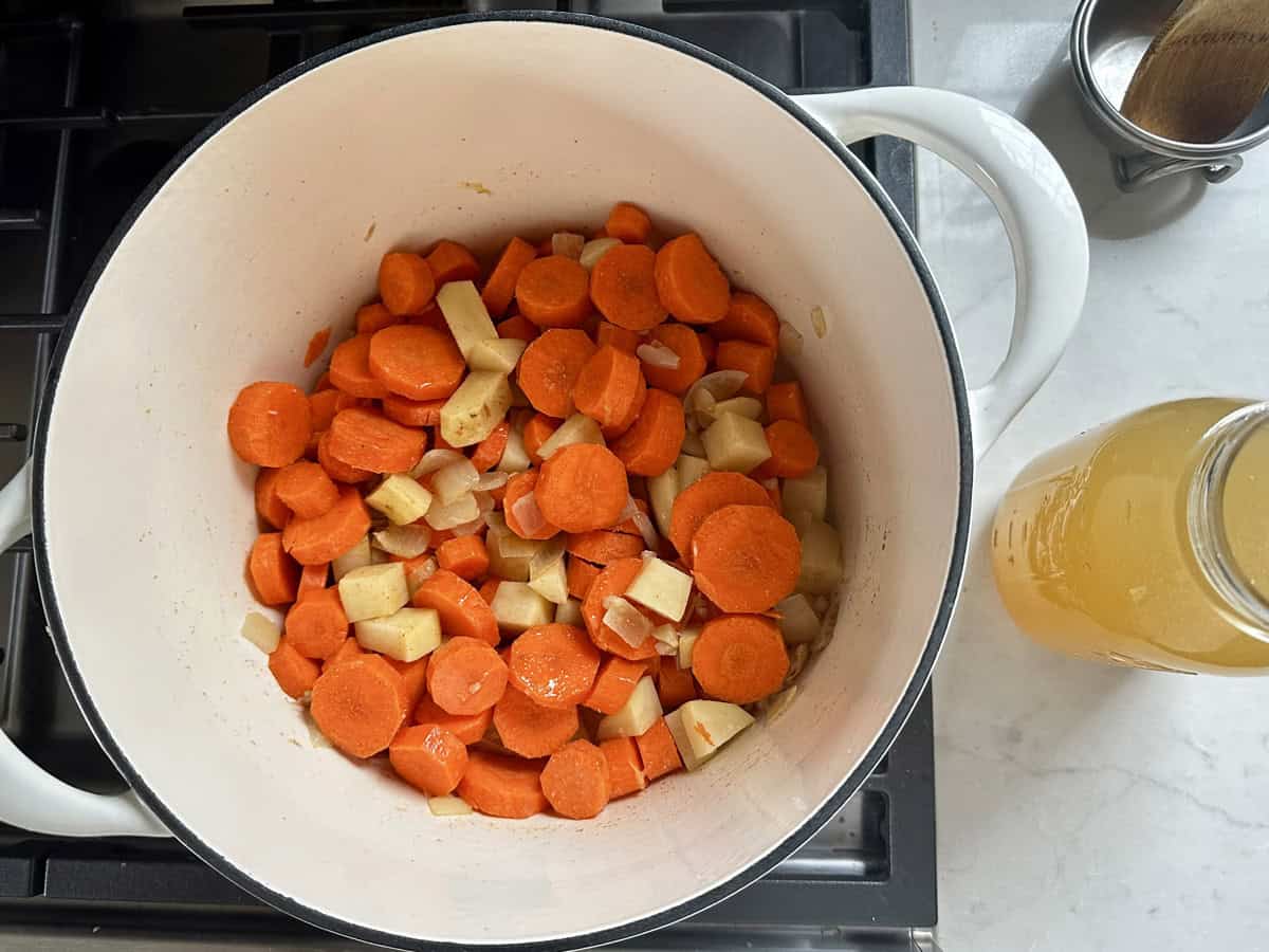 A white pot on a stove contains chopped carrots, parsnips, and onions. A jar of yellow vegetable broth sits nearby on a white countertop.