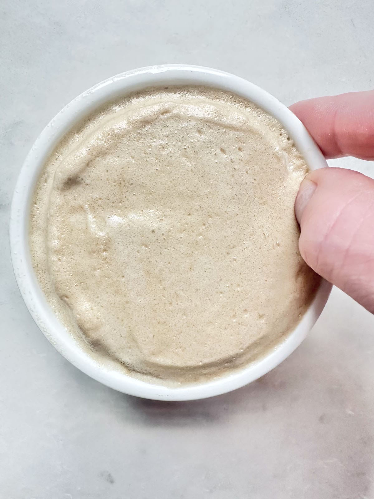 A hand holds a small white bowl filled with bubbly, creamy beige sourdough starter against a light, textured background.