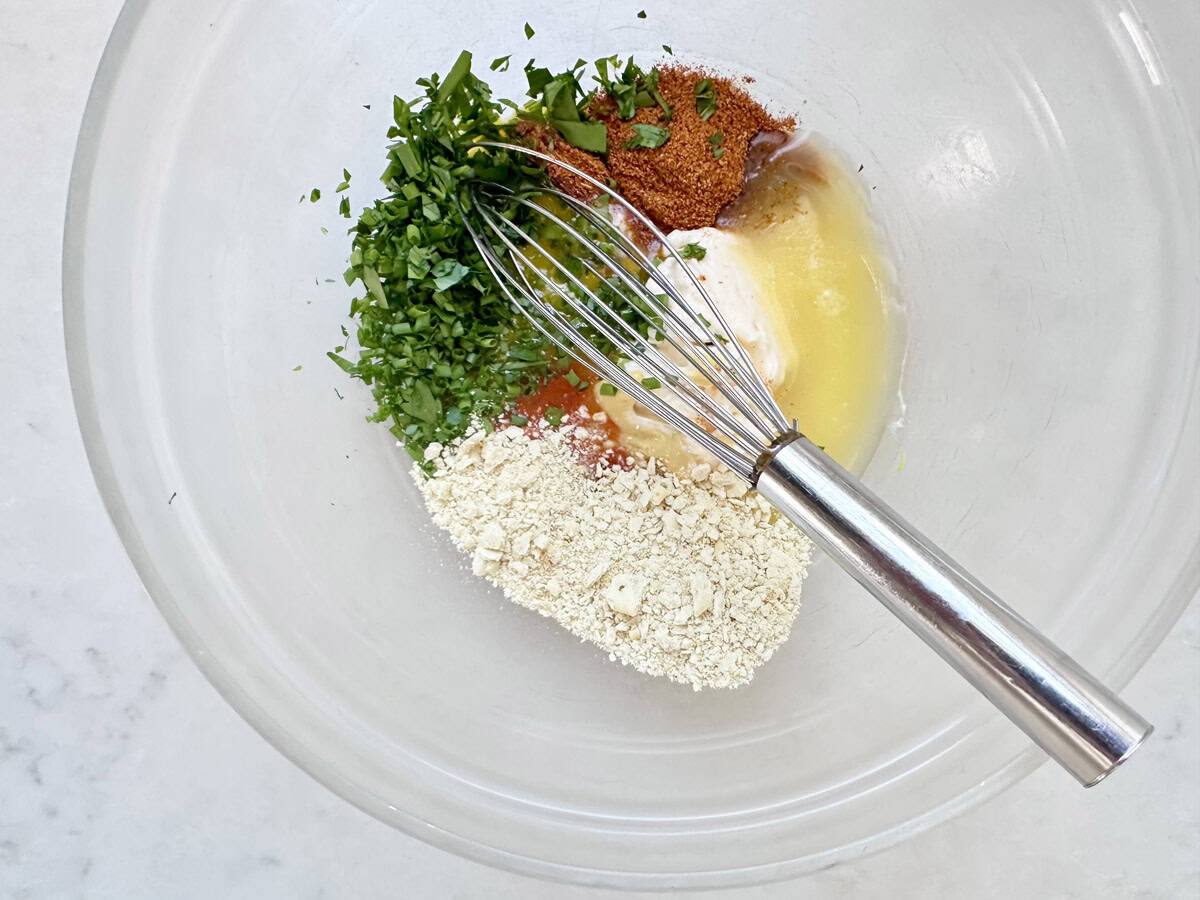 A clear mixing bowl contains chopped herbs, spices, oat flour, melted butter, and mayonnaise, with a metal whisk resting inside on a white surface.