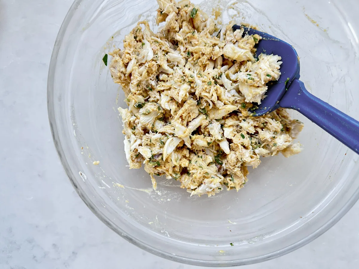 A glass bowl holds a mixture of shredded crab meat, herbs, and seasonings, being stirred with a blue spatula on a light-colored countertop.