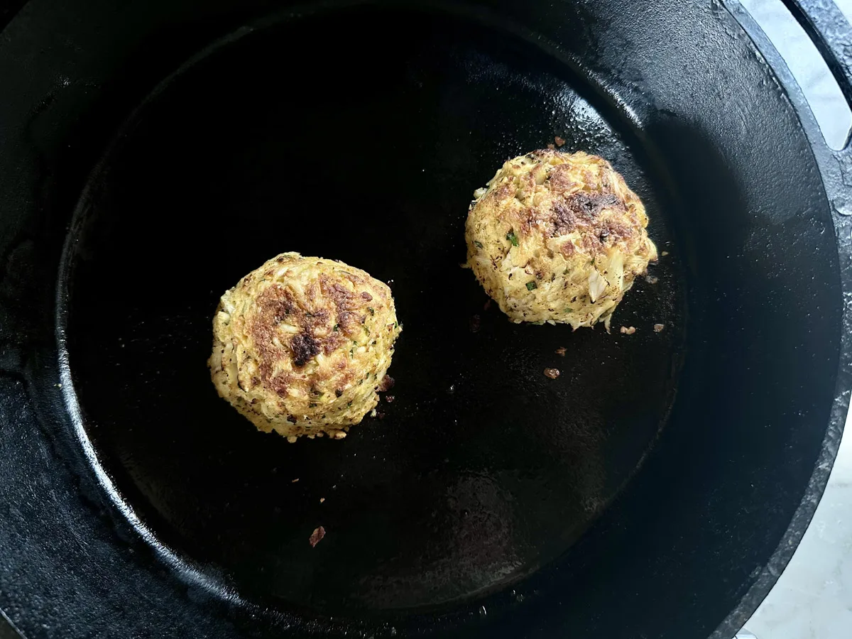 Two golden-brown patties are cooking in a black cast iron skillet, showing crisp, textured surfaces with slight browning, set against the dark background of the pan.