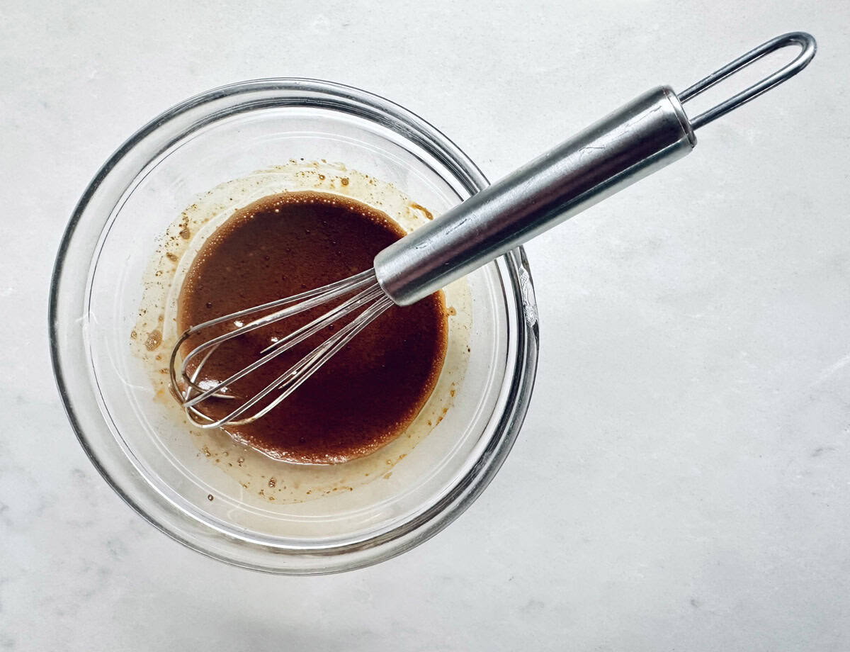 A metal whisk rests in a glass bowl containing a dark brown liquid mixture, set on a light-colored surface.