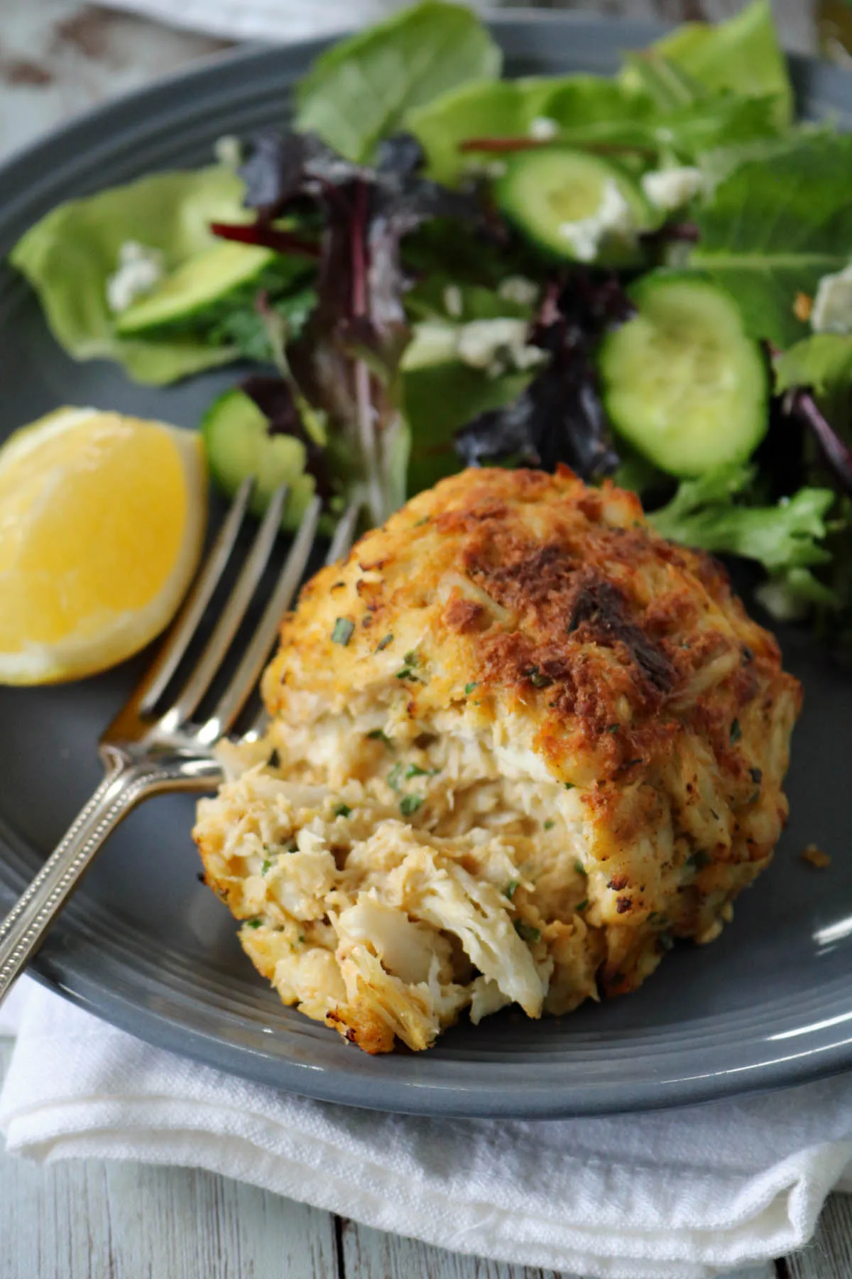 A crab cake served on a gray plate with a leafy green salad, cucumber slices, a lemon wedge, and a fork on the side.