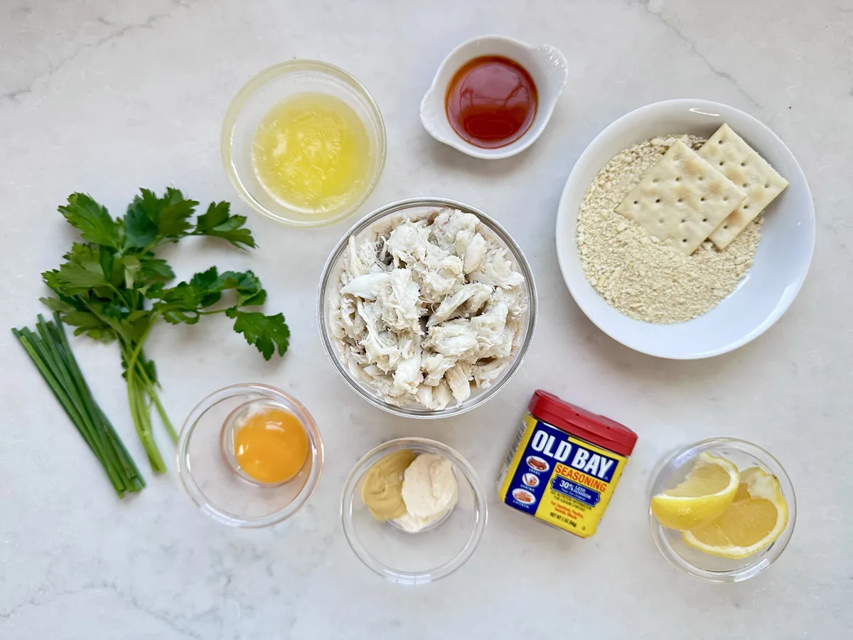A selection of ingredients for crab cakes: crab meat, egg yolk, melted butter, hot sauce, mayonnaise, mustard, Old Bay seasoning, parsley, chives, lemon wedges, oyster crackers, and saltine crackers on a white surface.