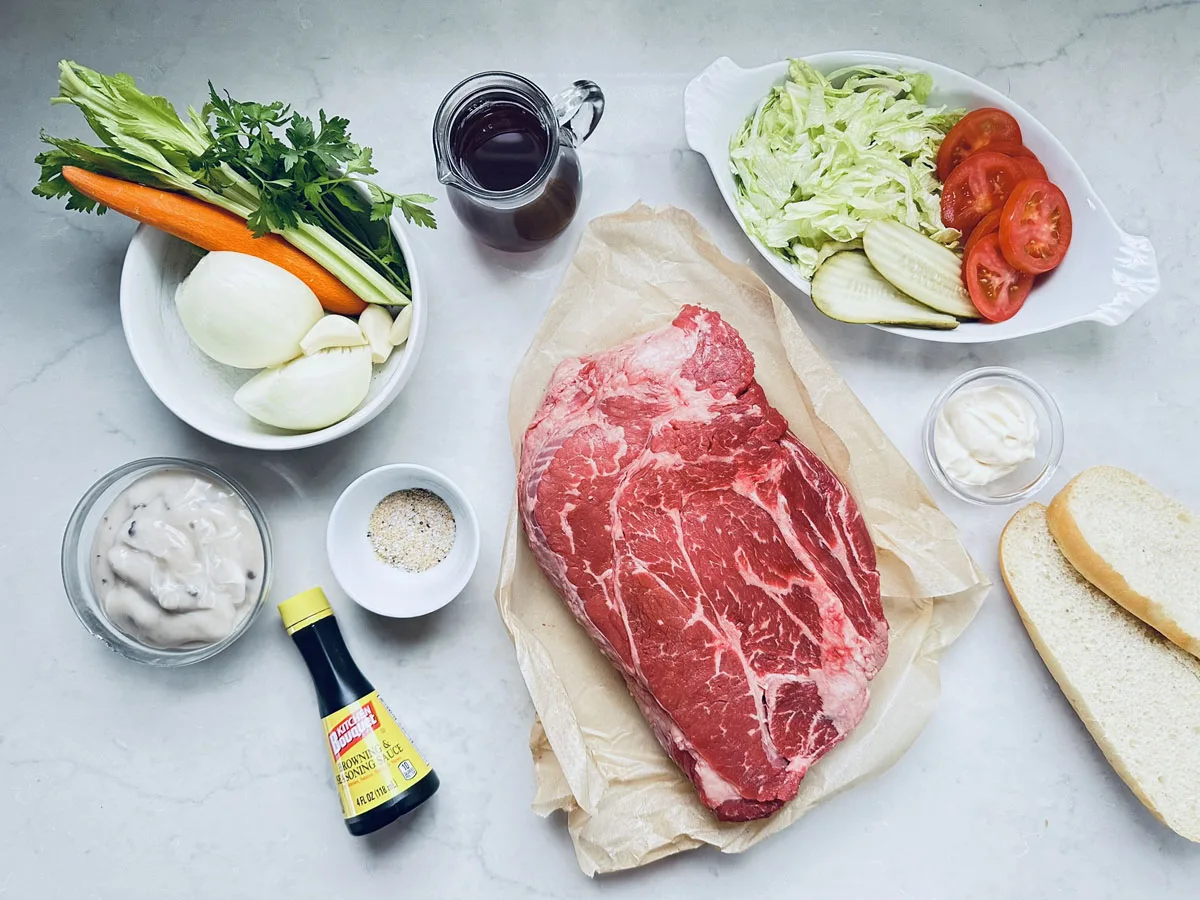 A slab of raw beef on parchment, surrounded by sliced onions, carrots, celery, lettuce, tomato, pickles, bread, mayonnaise, soup mix, seasoning, soy sauce, and a glass of dark liquid on a light countertop.