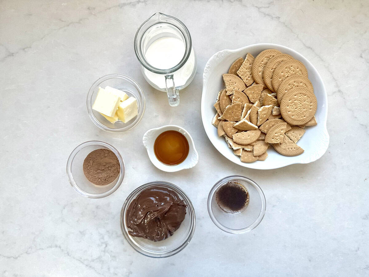 A top view of ingredients in bowls on a white surface: milk, butter, cocoa powder, vanilla extract, chocolate spread, a bowl of broken biscuits, and a small bowl of liquid, likely coffee.