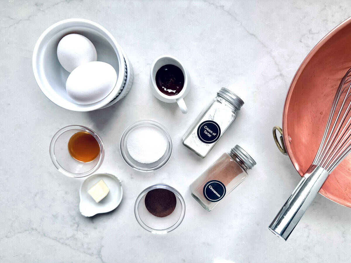 A top-down view of baking ingredients on a marble surface, including eggs, vanilla extract, sugar, butter, cocoa powder, chocolate, flour, cinnamon, and a copper mixing bowl with a whisk.