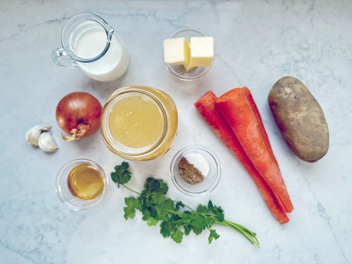 Top-down view of cooking ingredients on a white countertop, including carrots, a potato, an onion, garlic, fresh cilantro, butter, milk, broth, seasonings, and a small glass of vinegar.