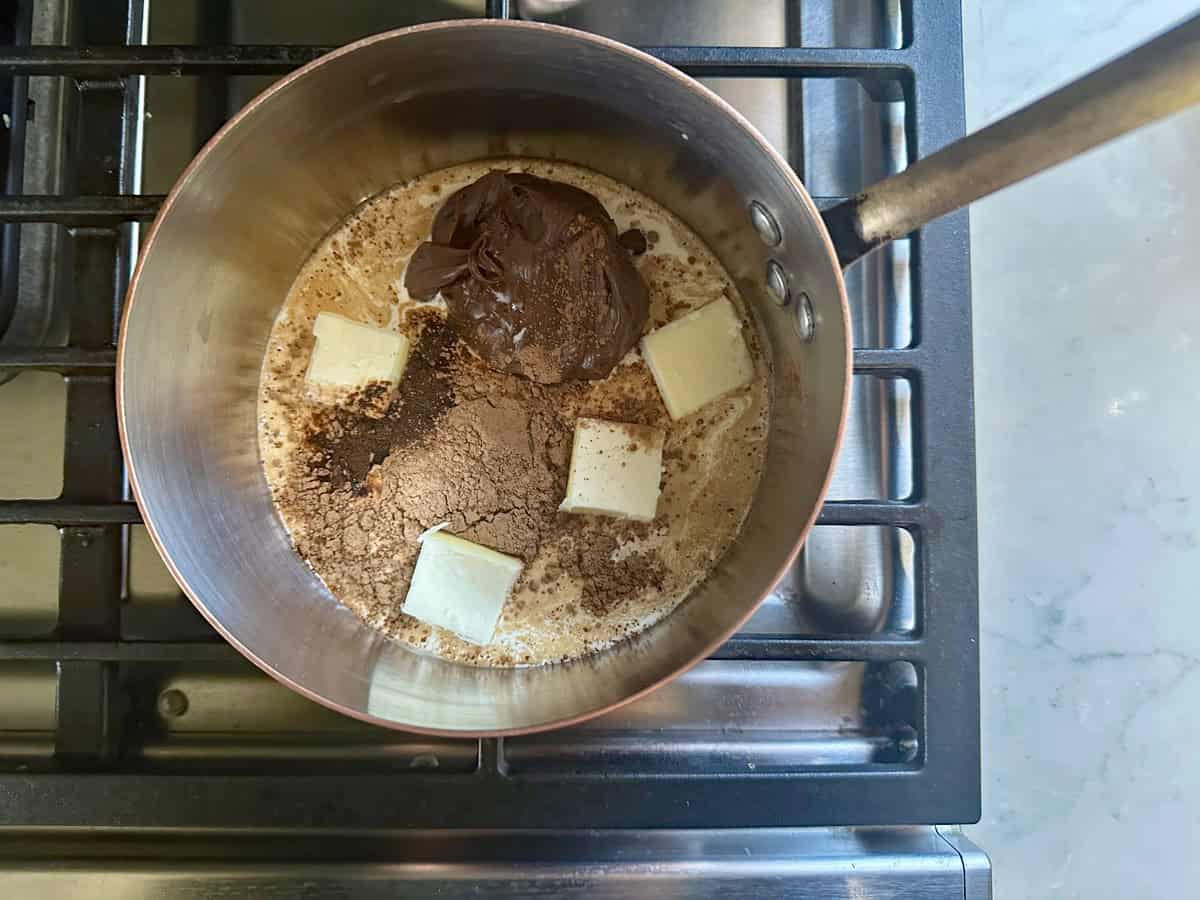 A saucepan on a stove contains melting butter, cocoa powder, and chocolate hazelnut spread, starting to blend together.