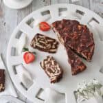 A white plate holds slices of chocolate biscuit cake, surrounded by fresh strawberries. A bowl of strawberries is nearby, and a knife rests on the table. The scene is set on a rustic wooden surface.