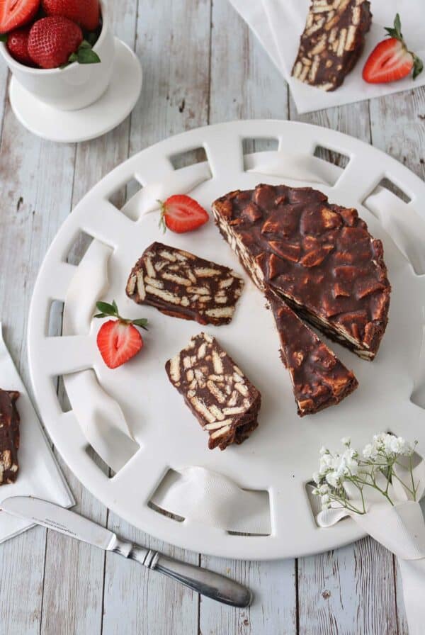 A white plate holds slices of chocolate biscuit cake, surrounded by fresh strawberries. A bowl of strawberries is nearby, and a knife rests on the table. The scene is set on a rustic wooden surface.