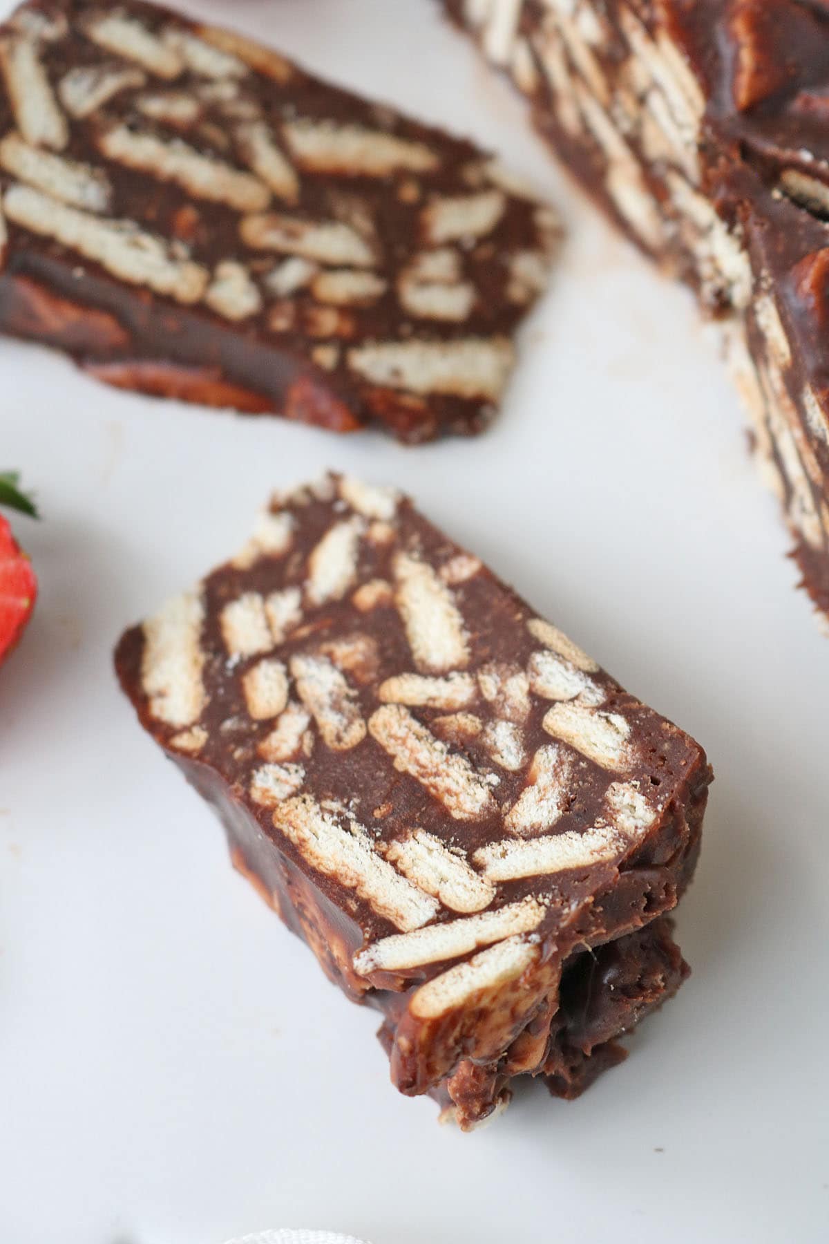 A close-up of a slice of chocolate biscuit cake featuring pieces of biscuit embedded in a rich, chocolate mixture, placed on a white surface with part of a strawberry visible nearby.