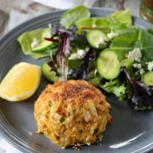 A crab cake sits on a gray plate next to a mixed green salad with cucumber slices and crumbled cheese, and a lemon wedge on the side. The plate rests on a white napkin on a rustic wooden table.