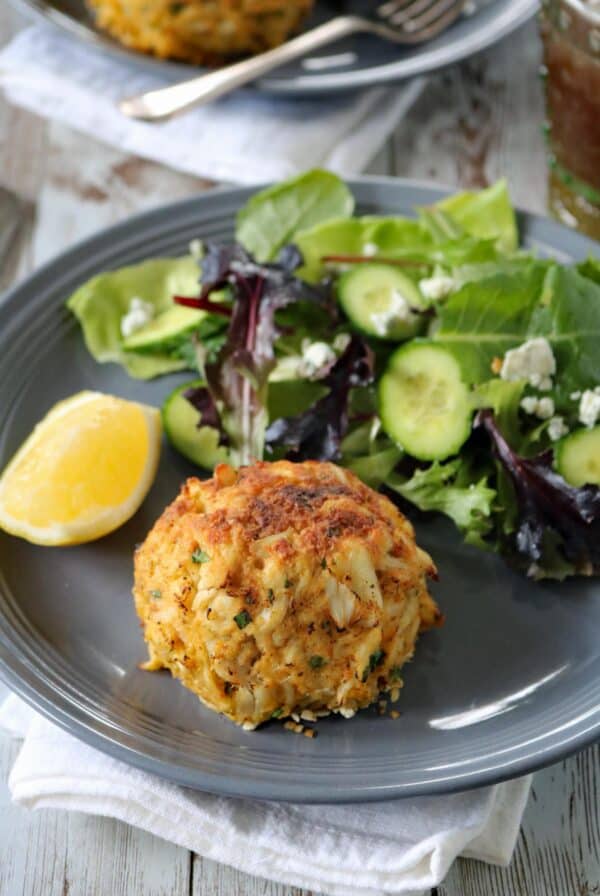 A crab cake sits on a gray plate next to a mixed green salad with cucumber slices and crumbled cheese, and a lemon wedge on the side. The plate rests on a white napkin on a rustic wooden table.