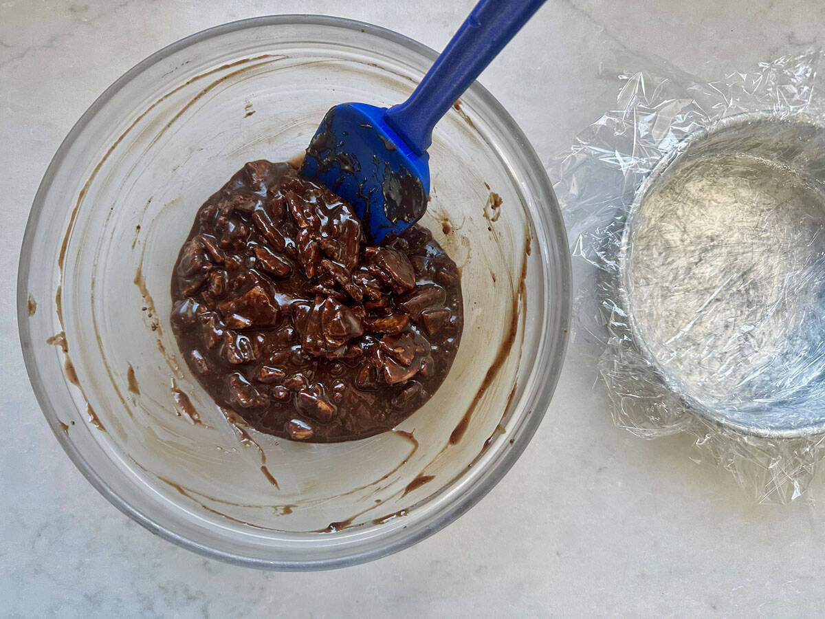 A glass bowl with chocolate and nuts mixture is being stirred with a blue spatula. Next to it is a smaller bowl lined with plastic wrap on a white countertop.