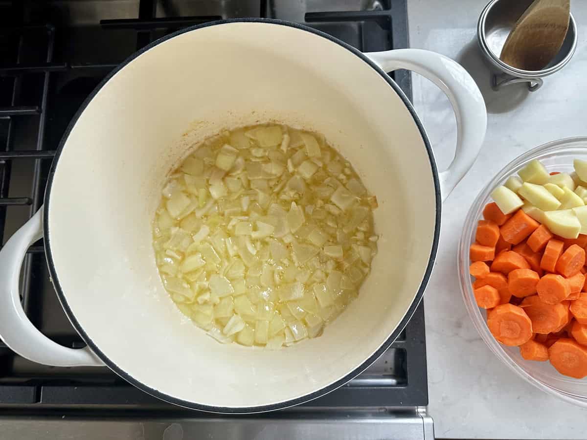 Chopped onions sautéing in a white pot on a stove, with a bowl of sliced carrots and diced potatoes next to it on a countertop.