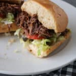 A shredded beef sandwich with lettuce, tomato, and pickles on a white plate, with a black and white checkered napkin beside it. The sandwich is cut in half, showing the filling.