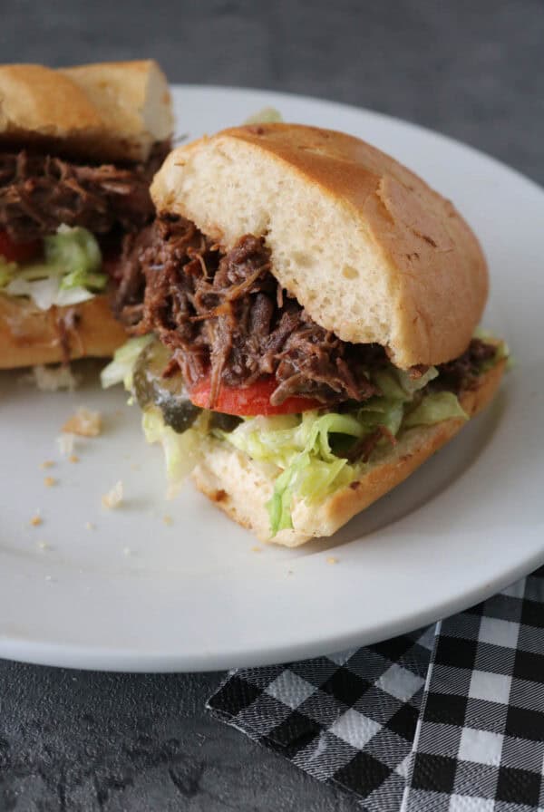 A shredded beef sandwich with lettuce, tomato, and pickles on a white plate, with a black and white checkered napkin beside it. The sandwich is cut in half, showing the filling.
