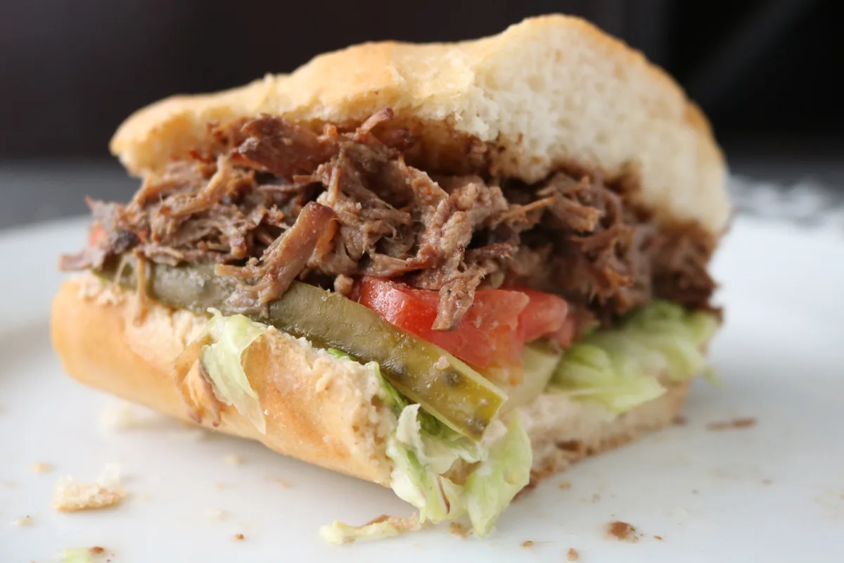 A close-up of a half-eaten sandwich on a white plate, filled with shredded beef, lettuce, tomato slices, pickles, and a soft sandwich bun.