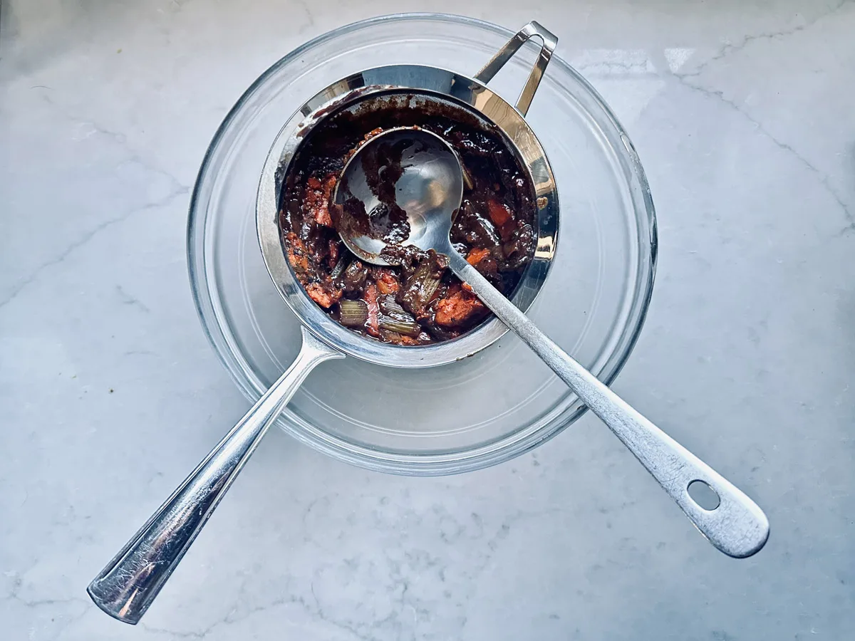 A metal strainer with a spoon rests over a clear glass bowl. The strainer contains chunky, dark red sauce with visible vegetable pieces, set on a light gray countertop.