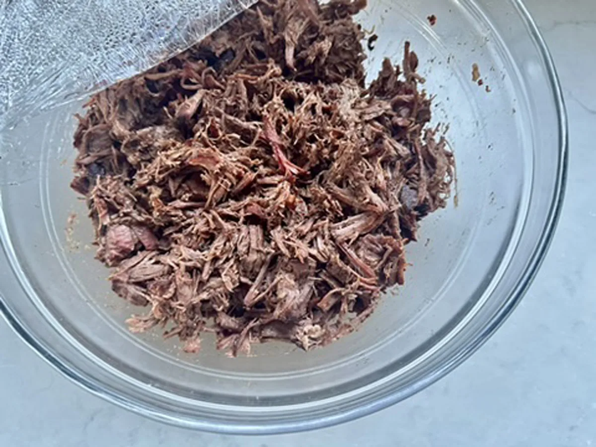A clear glass bowl filled with shredded cooked meat sits on a light-colored surface, partially covered with plastic wrap.