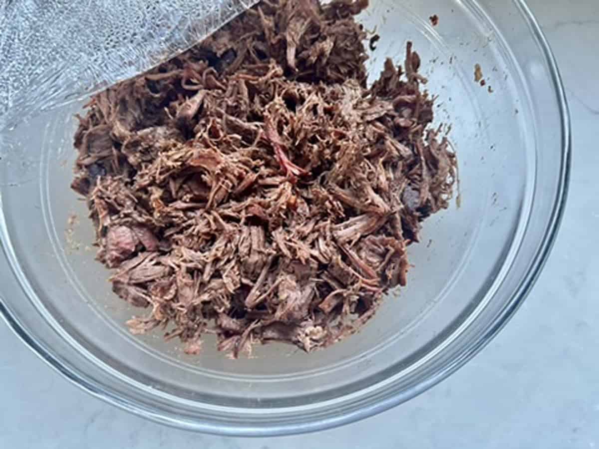 A clear glass bowl filled with shredded cooked meat sits on a light-colored surface, partially covered with plastic wrap.