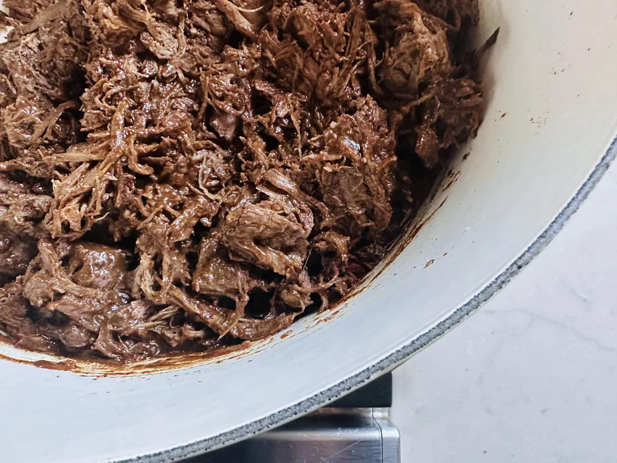 Shredded beef in a rich, dark sauce fills a white pot, with tender meat strands and chunks visible along the curved edge of the pot, set on a light-colored countertop.