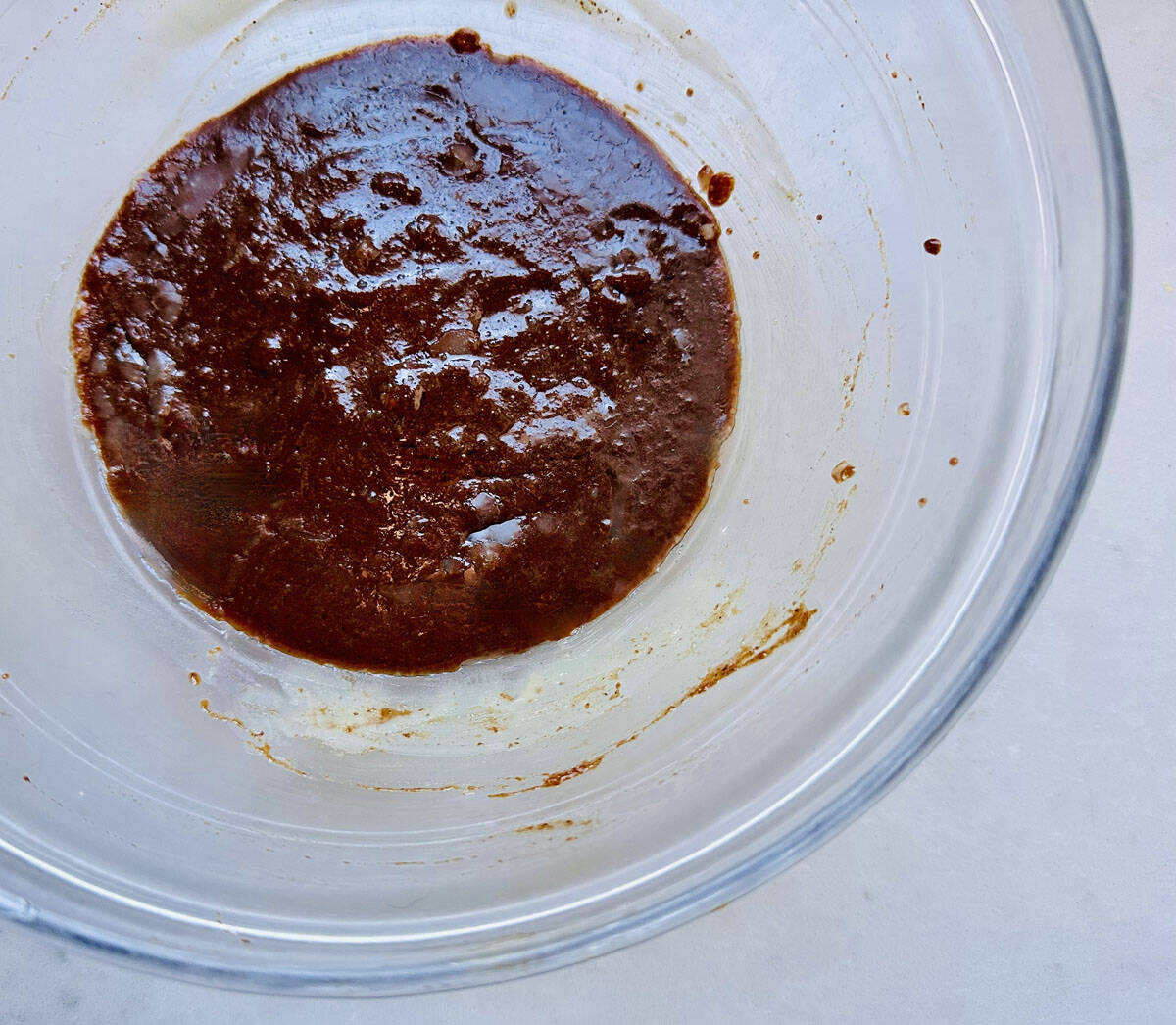 A clear glass bowl with a thick, dark brown mixture spread across the bottom and some residue on the sides, sitting on a light-colored surface.