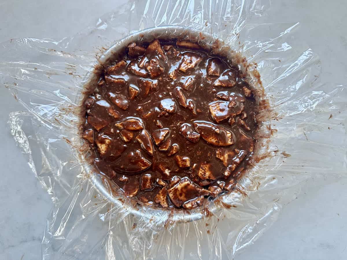 A round dish lined with plastic wrap is filled with a chocolate mixture containing visible chunks, sitting on a light-colored surface.
