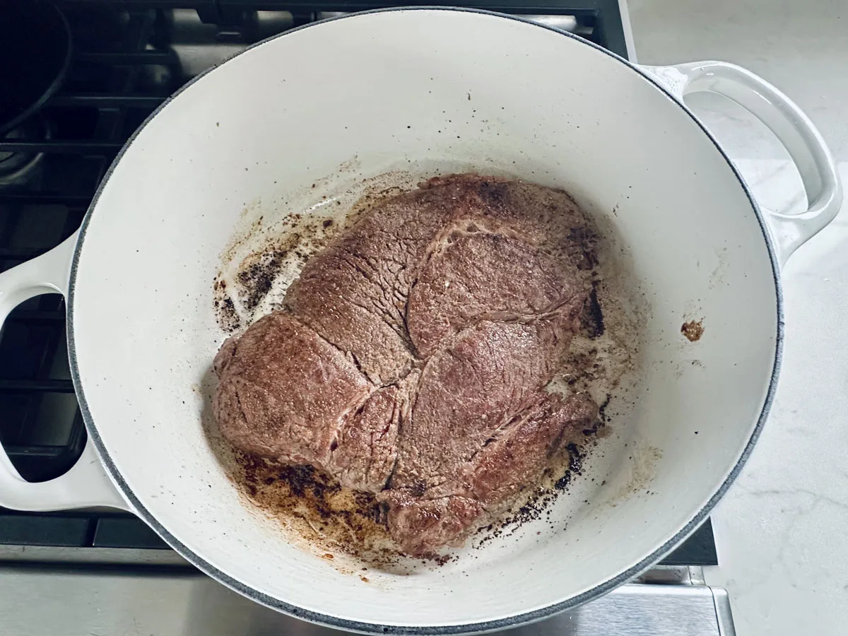 A large piece of beef is searing in a white Dutch oven on a stovetop, with browned edges and some seasoning visible.