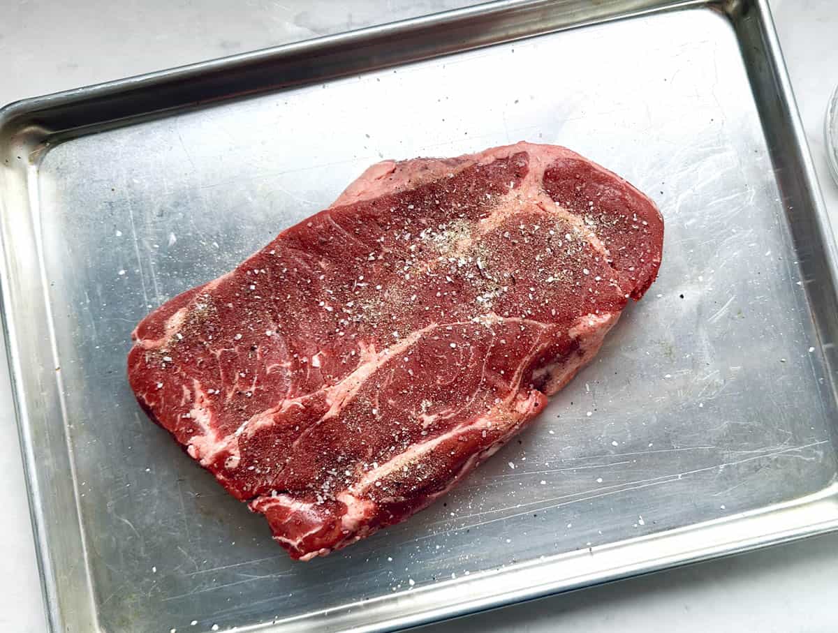 A raw, seasoned steak rests on a metal baking sheet. The steak has visible marbling and is sprinkled with salt and pepper. The baking sheet sits on a white surface.