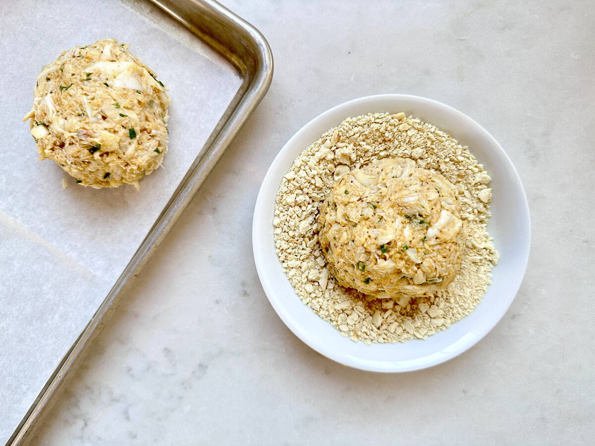 A ball of crab cake mixture sits in a shallow bowl of crushed crackers, while another coated crab cake rests on a parchment-lined baking sheet nearby on a light countertop.
