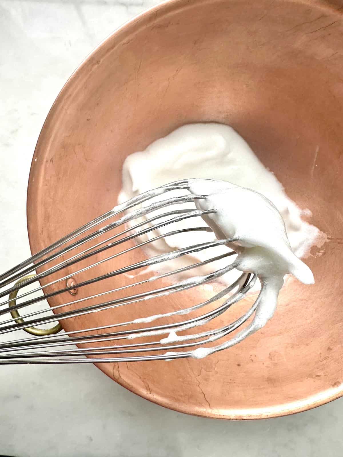 A metal whisk with soft white peaks of whipped egg whites rests over a copper mixing bowl containing more whipped egg whites, on a white countertop.