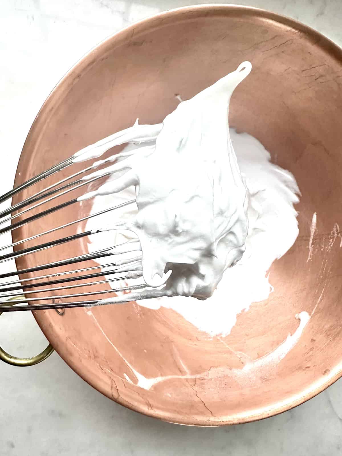 A metal whisk lifts glossy, stiff peaks of whipped egg whites from a copper mixing bowl on a light countertop.