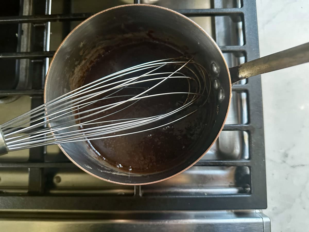 A metal whisk rests inside a small saucepan containing a dark liquid, sitting on a stovetop with black grates and a light-colored countertop visible beside it.