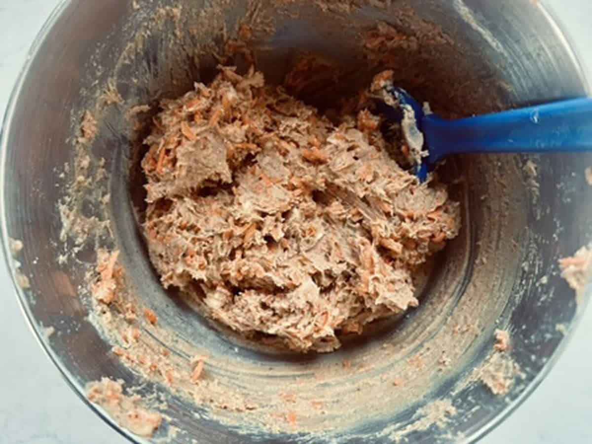 A metal mixing bowl filled with thick, textured batter containing visible shredded carrots, being stirred with a blue spatula.