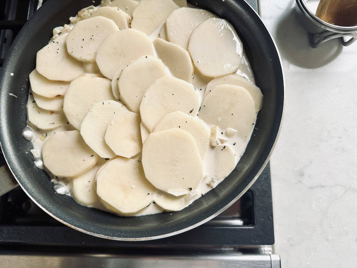 Sliced potatoes cooking in a creamy liquid in a black skillet on a stovetop, with bubbles forming around the edges. A small metal container is seen on the white countertop nearby.