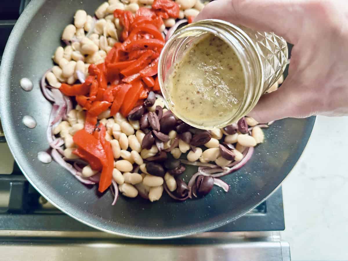 A hand pours dressing from a jar into a skillet containing white beans, sliced red peppers, olives, red onions, and other arugula salad ingredients on a stove—a fresh take on quick salad meals.