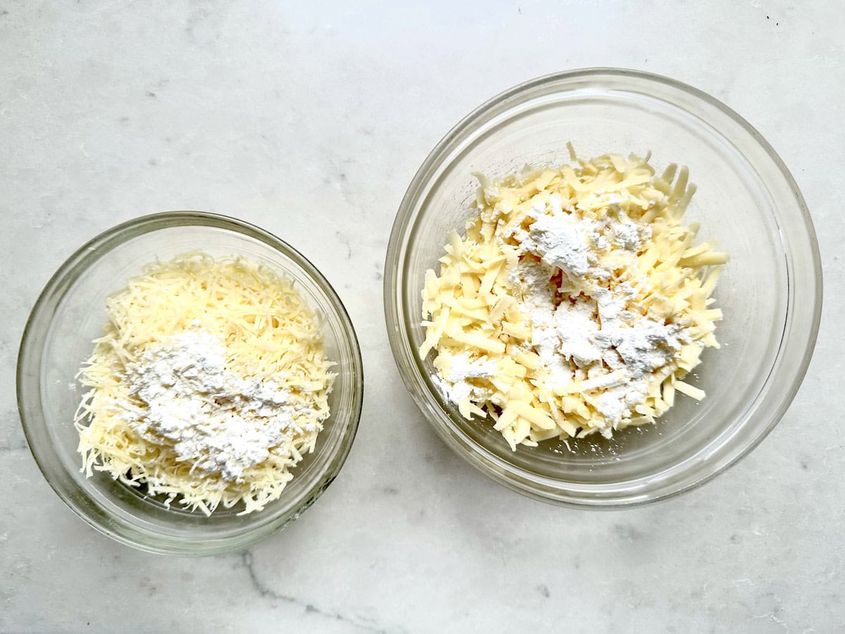 Two glass bowls on a light surface, each filled with grated cheese topped with a small amount of white flour. The bowl on the right is larger and contains more cheese than the smaller bowl on the left.