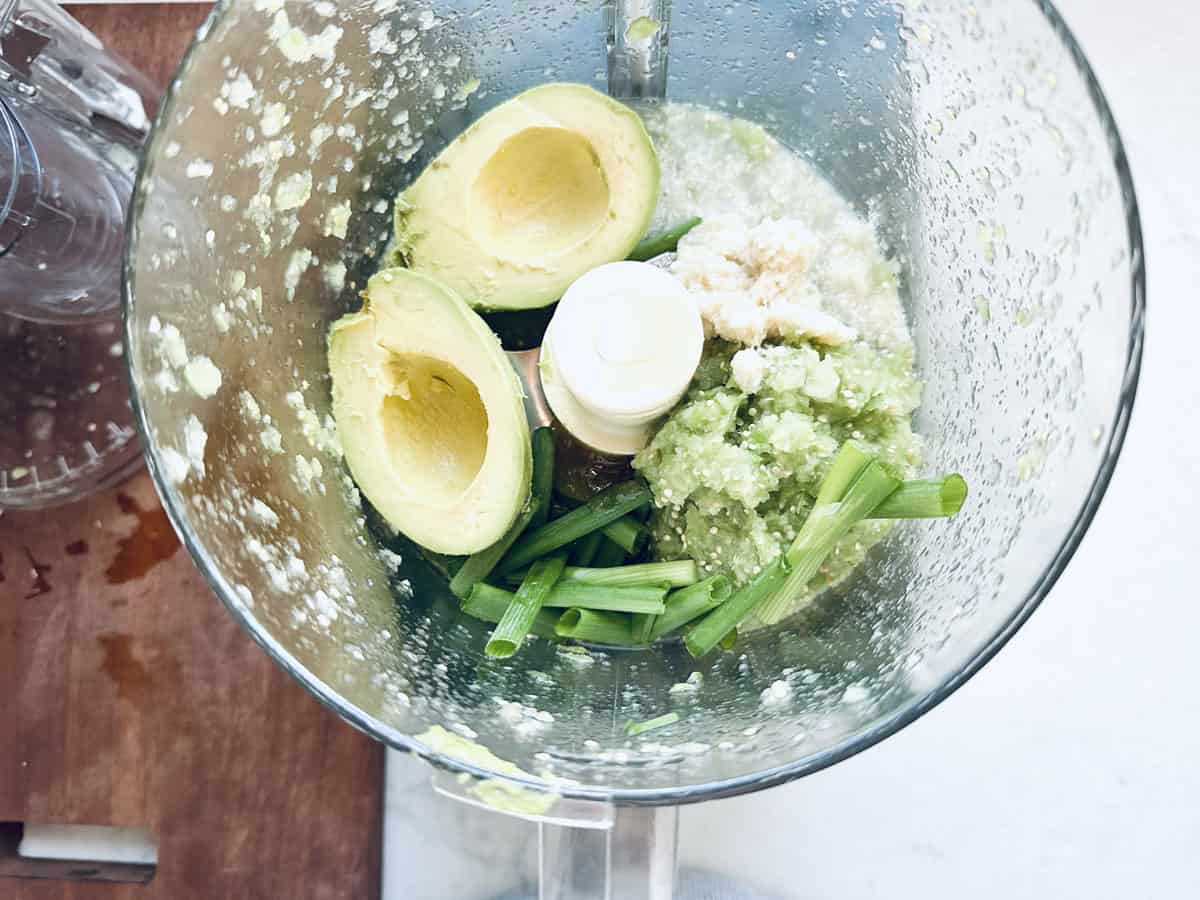 A food processor bowl containing halved avocado, chopped green onions, minced garlic, and a light green, blended mixture, viewed from above on a kitchen counter.