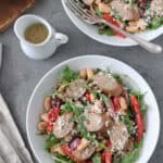 Two white bowls filled with salad featuring sliced sausage, white beans, arugula, red bell pepper, and grated cheese. A fork rests in one bowl. Nearby are sliced bread, a small jug of dressing, and napkins with cutlery.