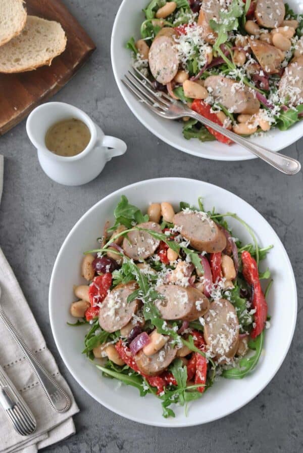 Two white bowls filled with salad featuring sliced sausage, white beans, arugula, red bell pepper, and grated cheese. A fork rests in one bowl. Nearby are sliced bread, a small jug of dressing, and napkins with cutlery.
