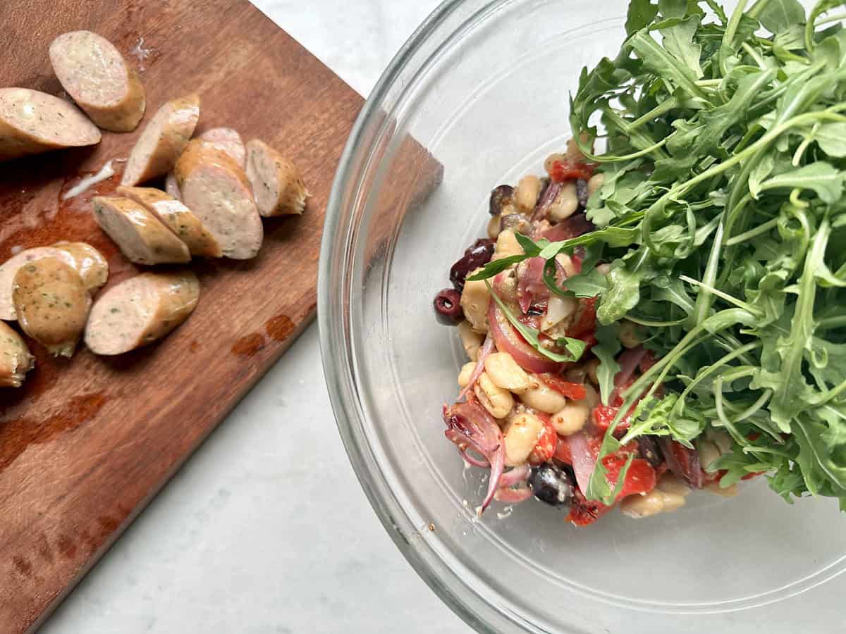 A glass bowl with arugula salad ingredients like beans, olives, and tomatoes, sits next to a wooden board with sliced sausage—perfect for sausage salad meals or an Arugula Salad with Sausage and White Beans.
